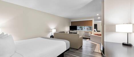 A hotel room with a large bed, a desk, a chair, a TV, and a kitchenette.