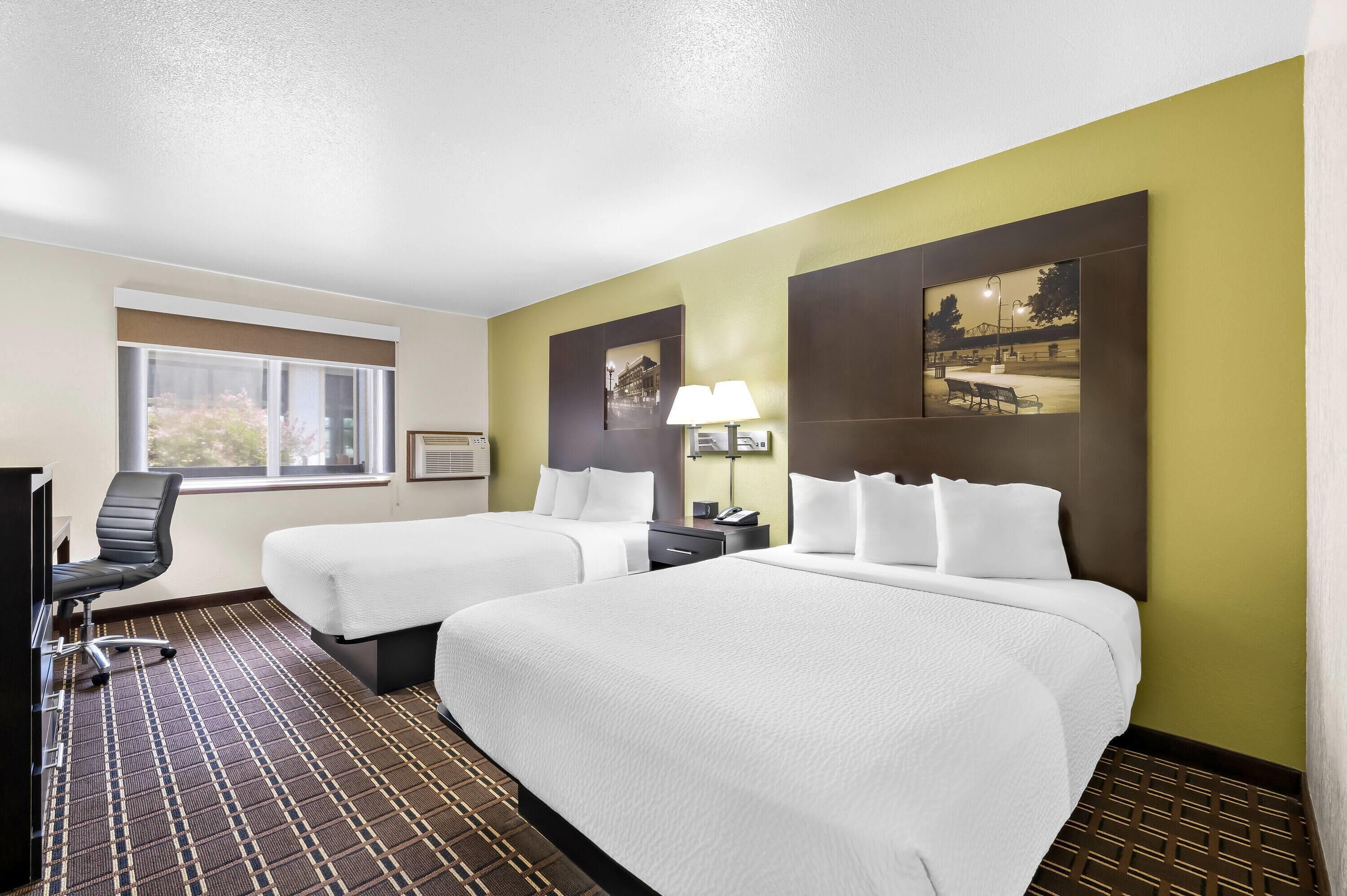 A hotel room with two beds, a desk, a chair, a lamp, and a window.