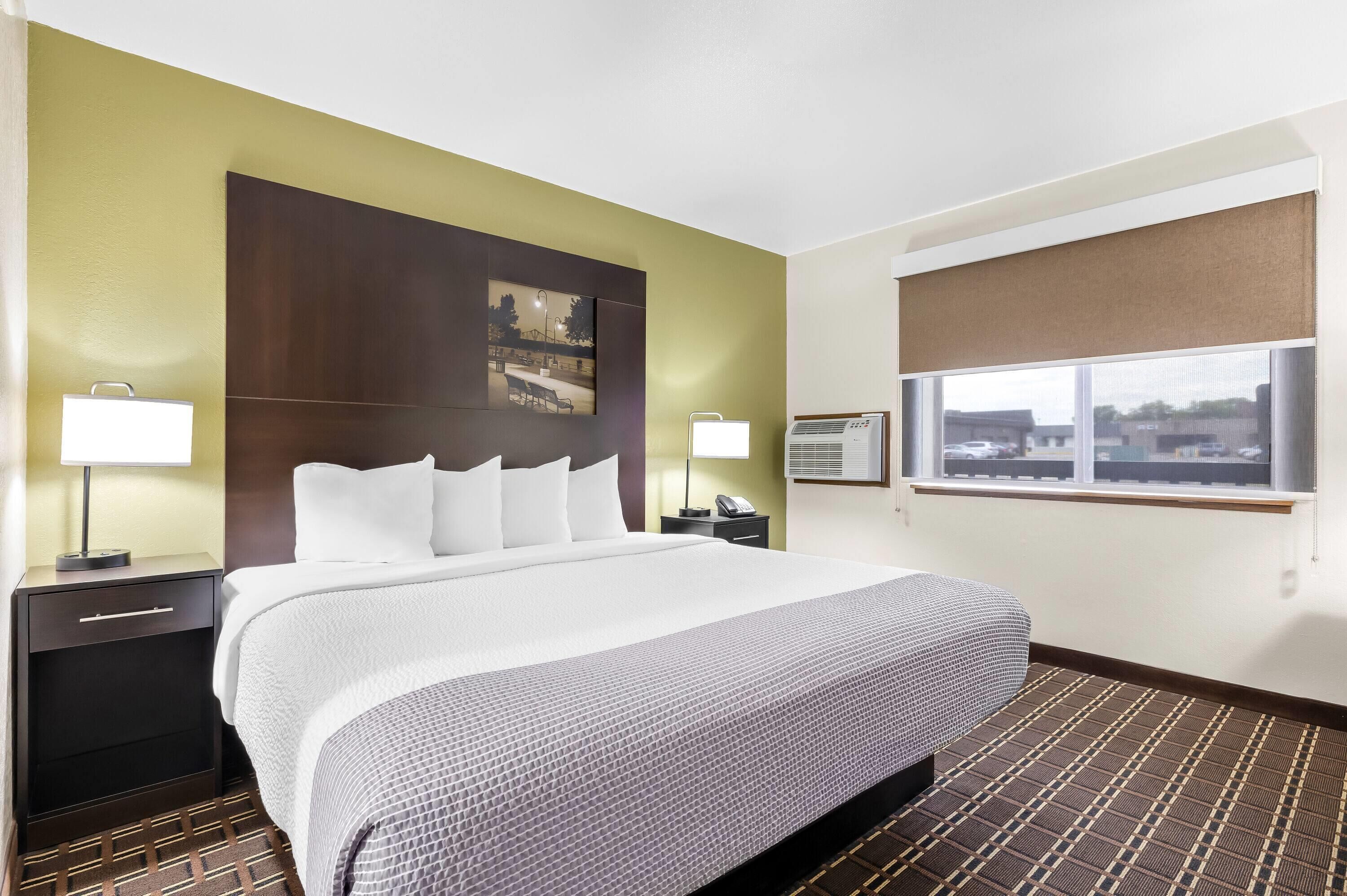 A hotel room with a large bed, two bedside tables with lamps, a wall-mounted air conditioner, and a window with a view of the city.