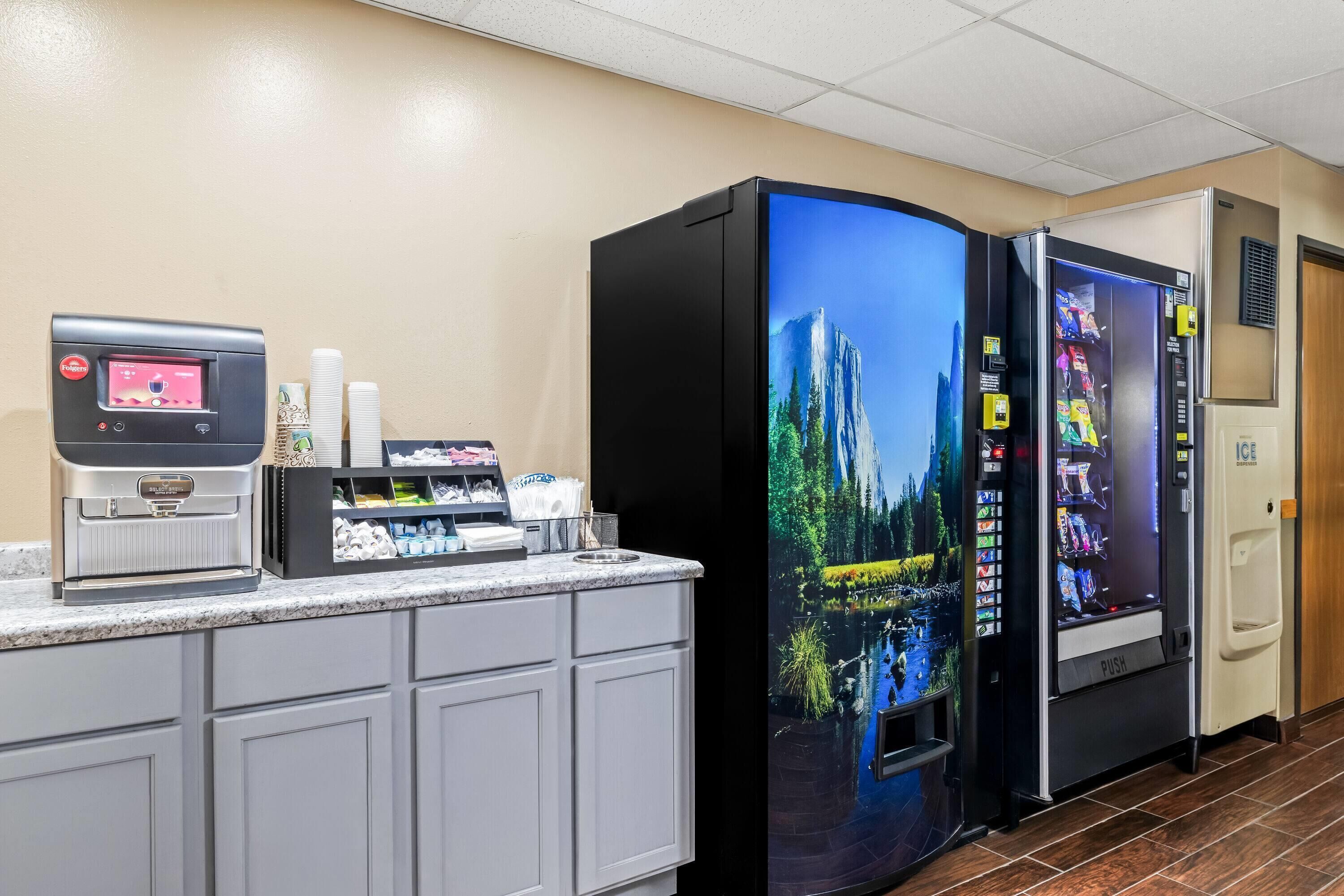 Una cocina de hotel con una máquina de café, un refrigerador de exhibición tipo cascada y una máquina expendedora.