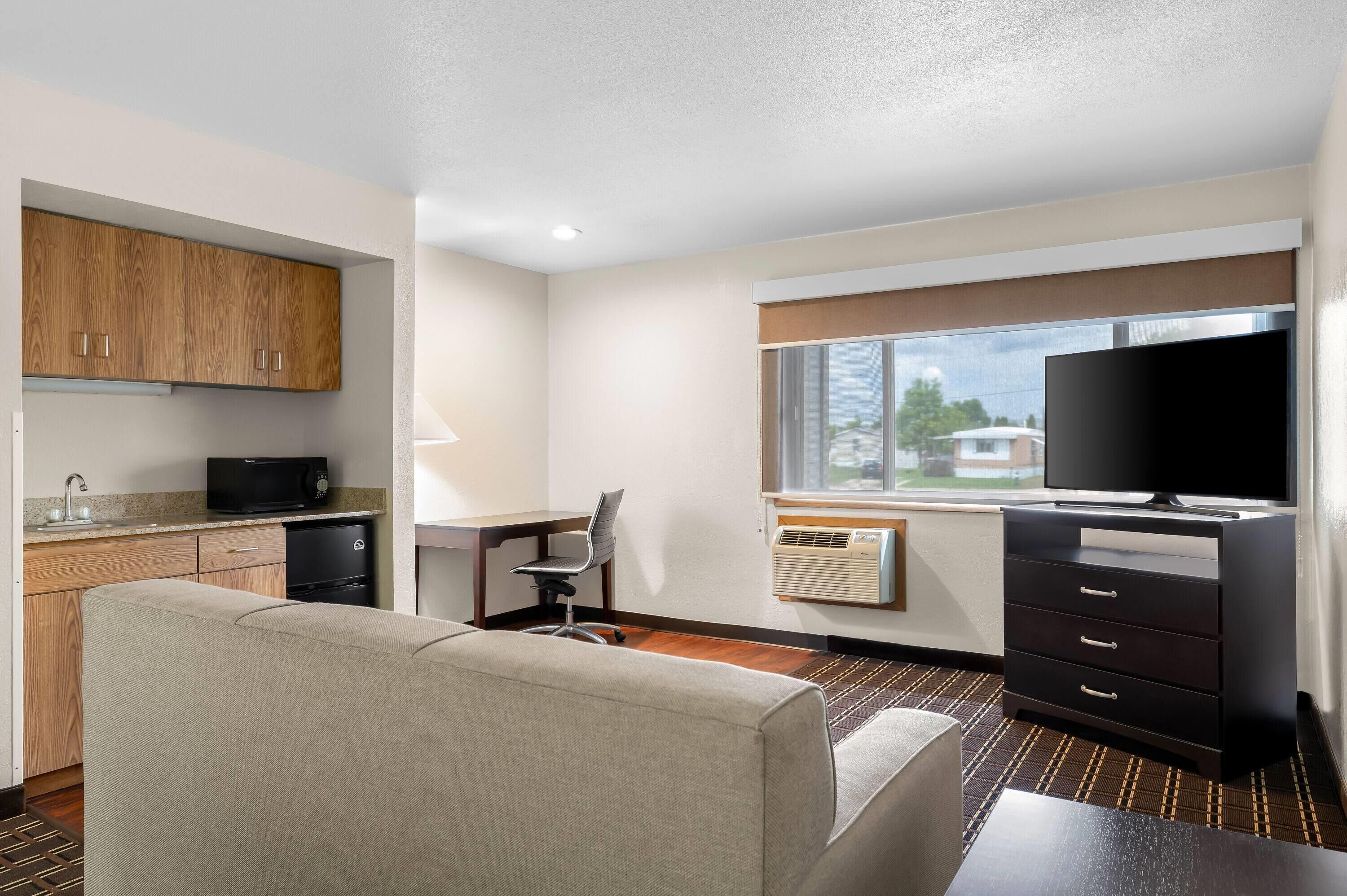A compact hotel room with a kitchenette, a sofa, a desk with a chair, a TV, and a window with a view of houses.