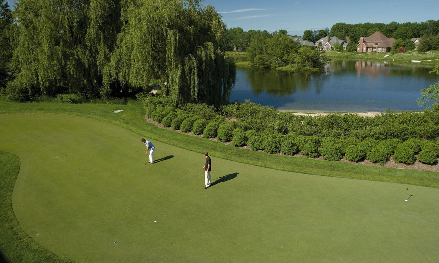 Un campo de golf con un estanque, árboles y casas al fondo.
