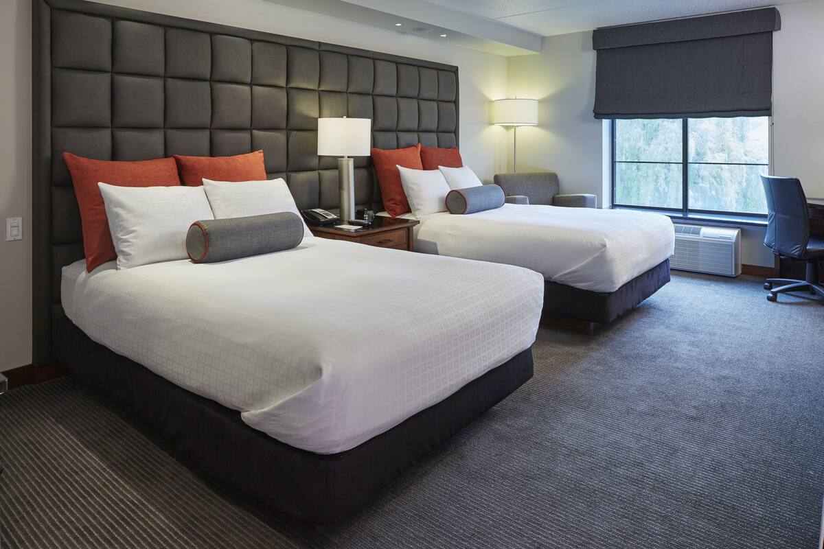 Habitación de hotel con dos camas, un cabecero grande y acolchado, un escritorio con una silla y una ventana con vistas a árboles.
