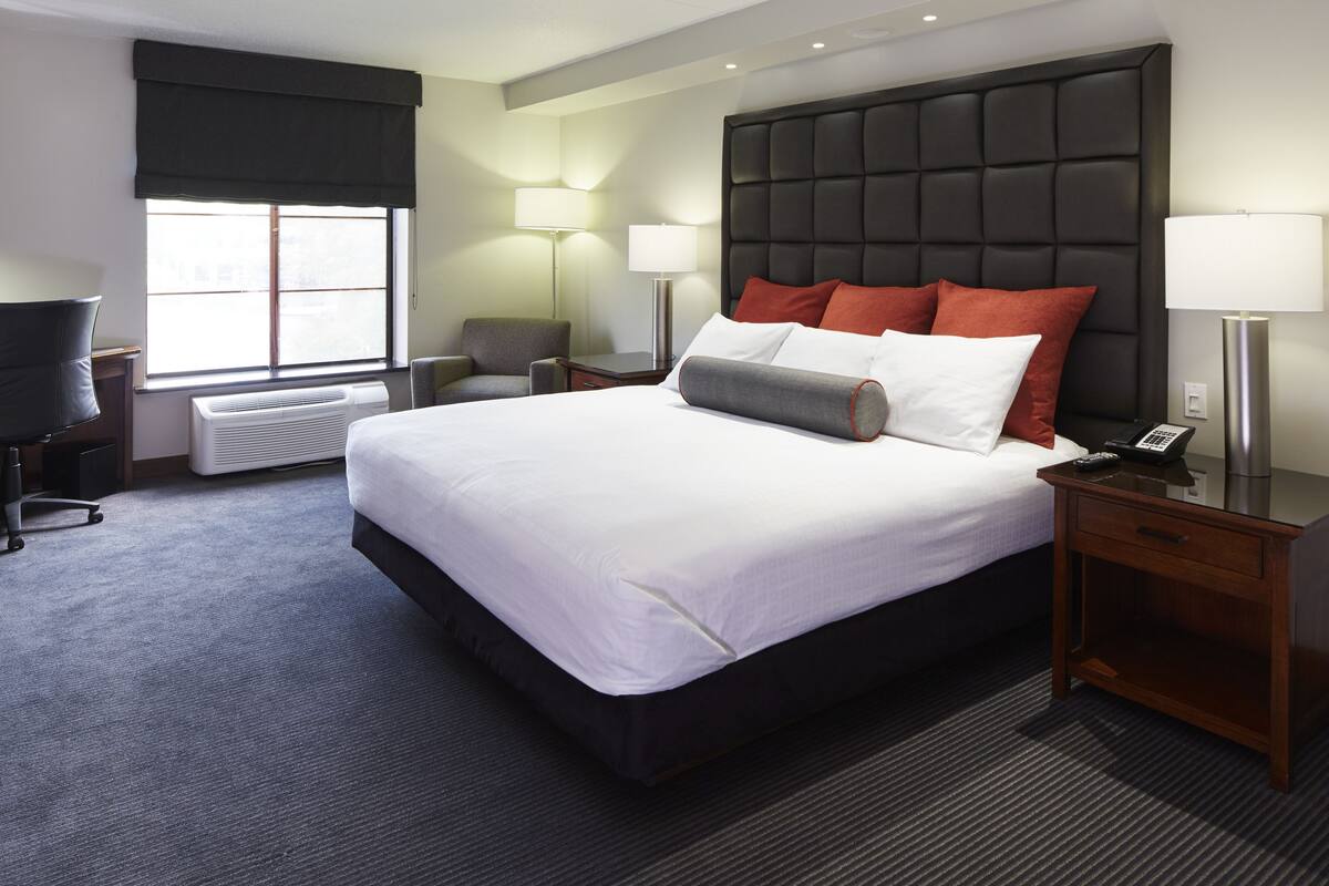Habitación de hotel con una cama grande, dos mesitas de noche, un escritorio, una silla, una ventana con persianas y un calentador de pared.