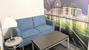A room with a blue sofa, a small round table with a lamp, and a large wall mural of fireworks and buildings. - AmericInn by Wyndham Green Bay Near Stadium (Green Bay)