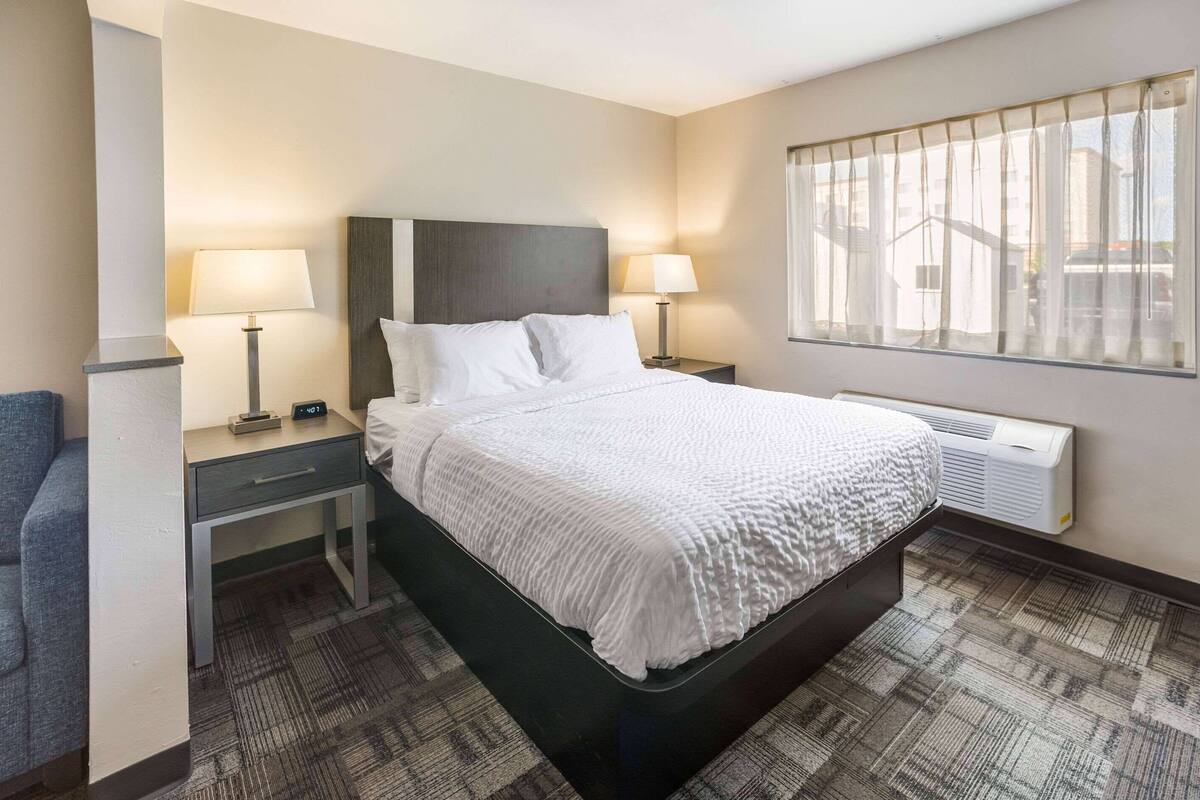 Habitación de hotel con una cama grande, dos mesitas de noche, un escritorio, una silla, una ventana con persianas y aire acondicionado de pared.