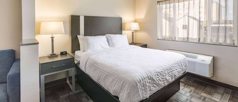 A hotel room with a large bed, two bedside tables, a desk, a chair, a window with blinds, and a wall-mounted air conditioner.