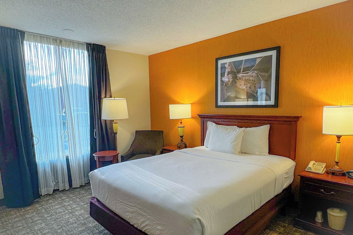 A hotel room with a large bed, two bedside tables with lamps, a chair, a painting on the wall, and a window with curtains.