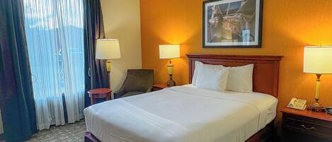 A hotel room with a large bed, two bedside tables with lamps, a chair, a painting on the wall, and a window with curtains.