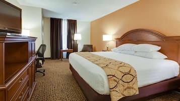 A hotel room with a large bed, a wooden dresser, a desk with a chair, a TV, and a window with curtains.