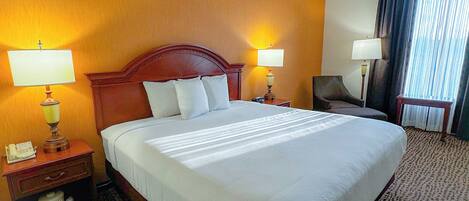A hotel room with a large bed, two bedside lamps, a chair, a desk with a telephone, and a window with curtains.