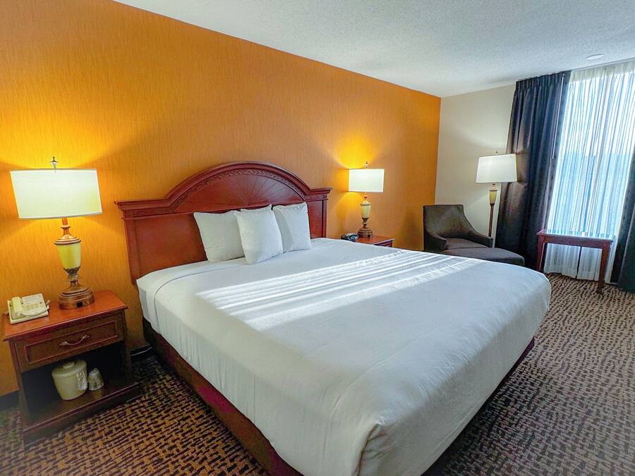 A hotel room with a large bed, two bedside lamps, a chair, a desk with a telephone, and a window with curtains.