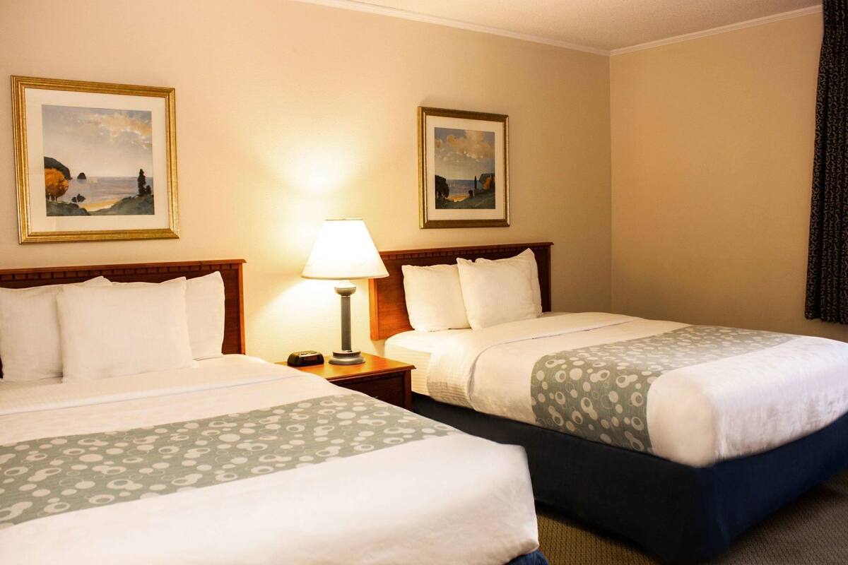 A hotel room with two beds, a nightstand with a lamp, and two framed landscape paintings on the wall.