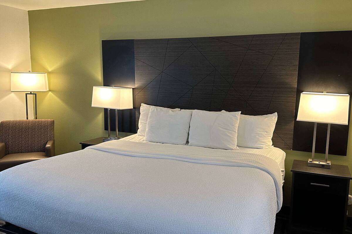 A neatly made bed with white bedding, two bedside lamps, a chair, and a dark headboard.