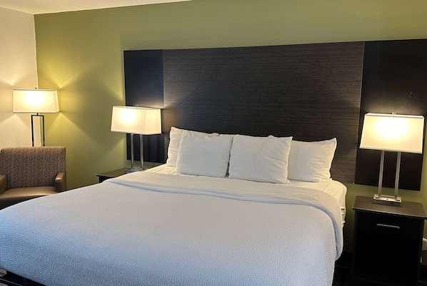 A neatly made bed with white bedding, two bedside lamps, a chair, and a dark headboard.