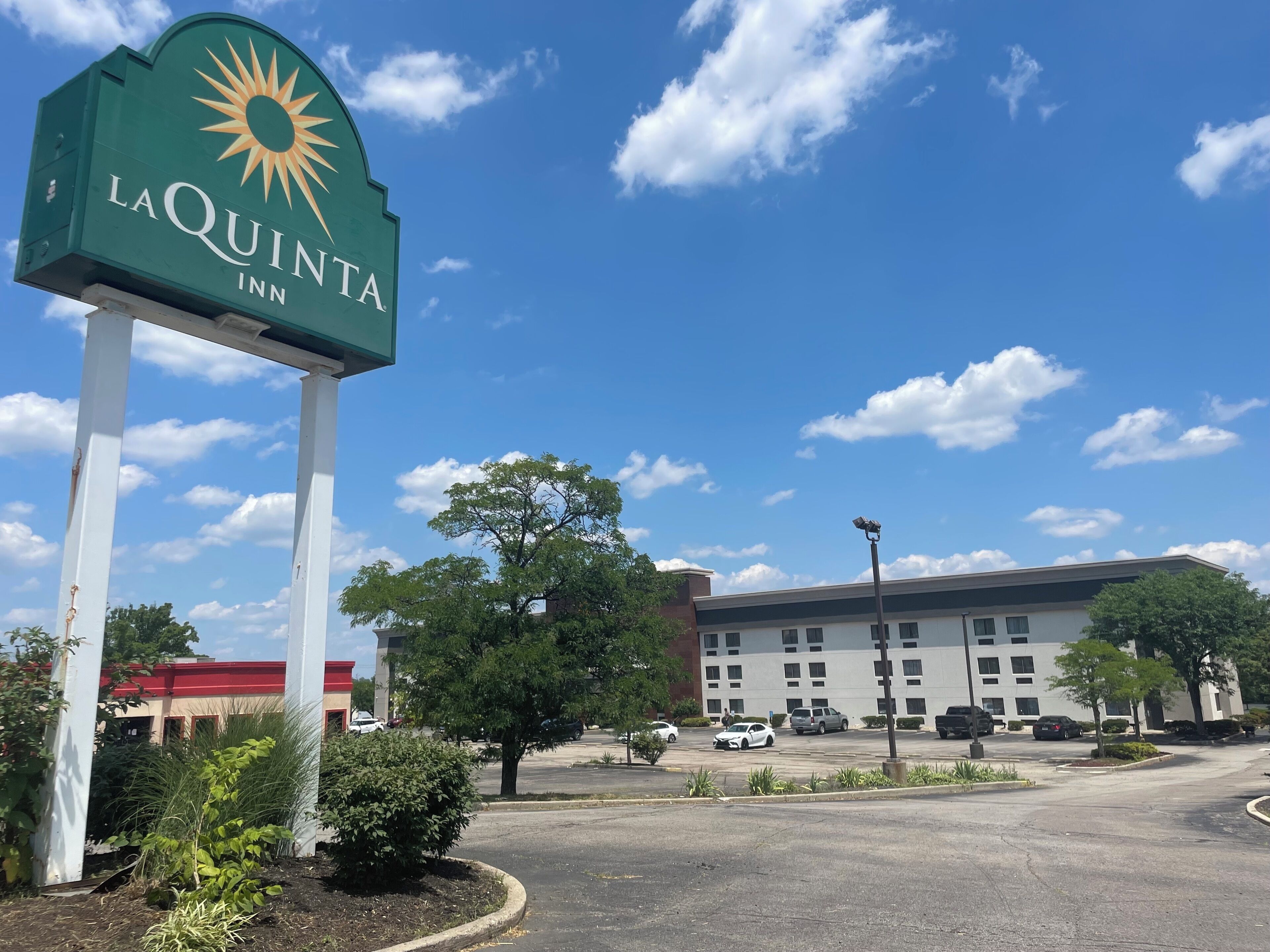 A La Quinta Inn sign with a sun emblem, surrounded by greenery and a parking lot with cars.