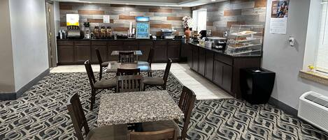 Habitación de hotel con comedor que incluye una mesa y sillas, una cocina con cafetera y refrigerador, y una alfombra estampada.