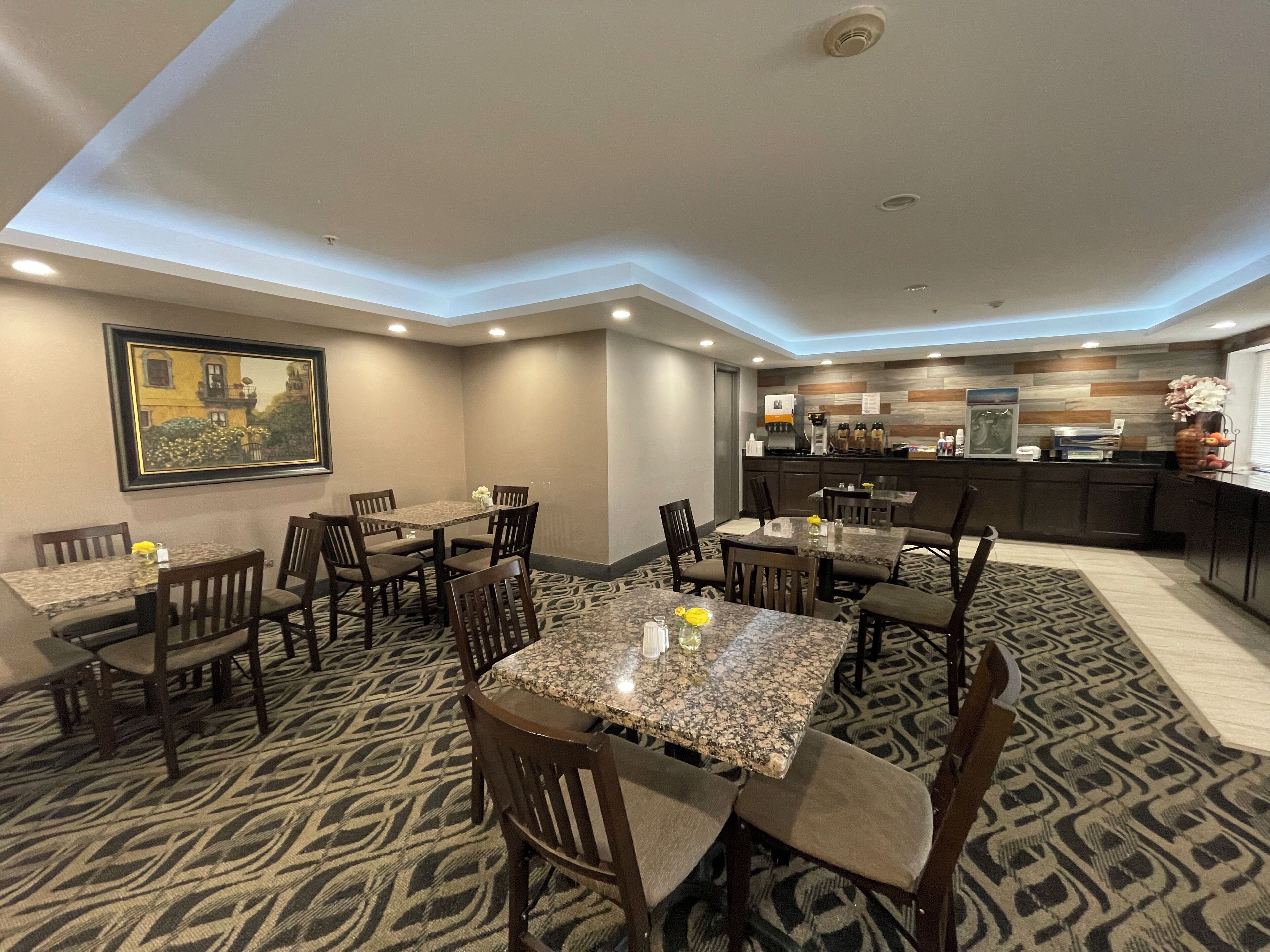 Un comedor de hotel con varias mesas y sillas, una cocina al fondo y una alfombra estampada.