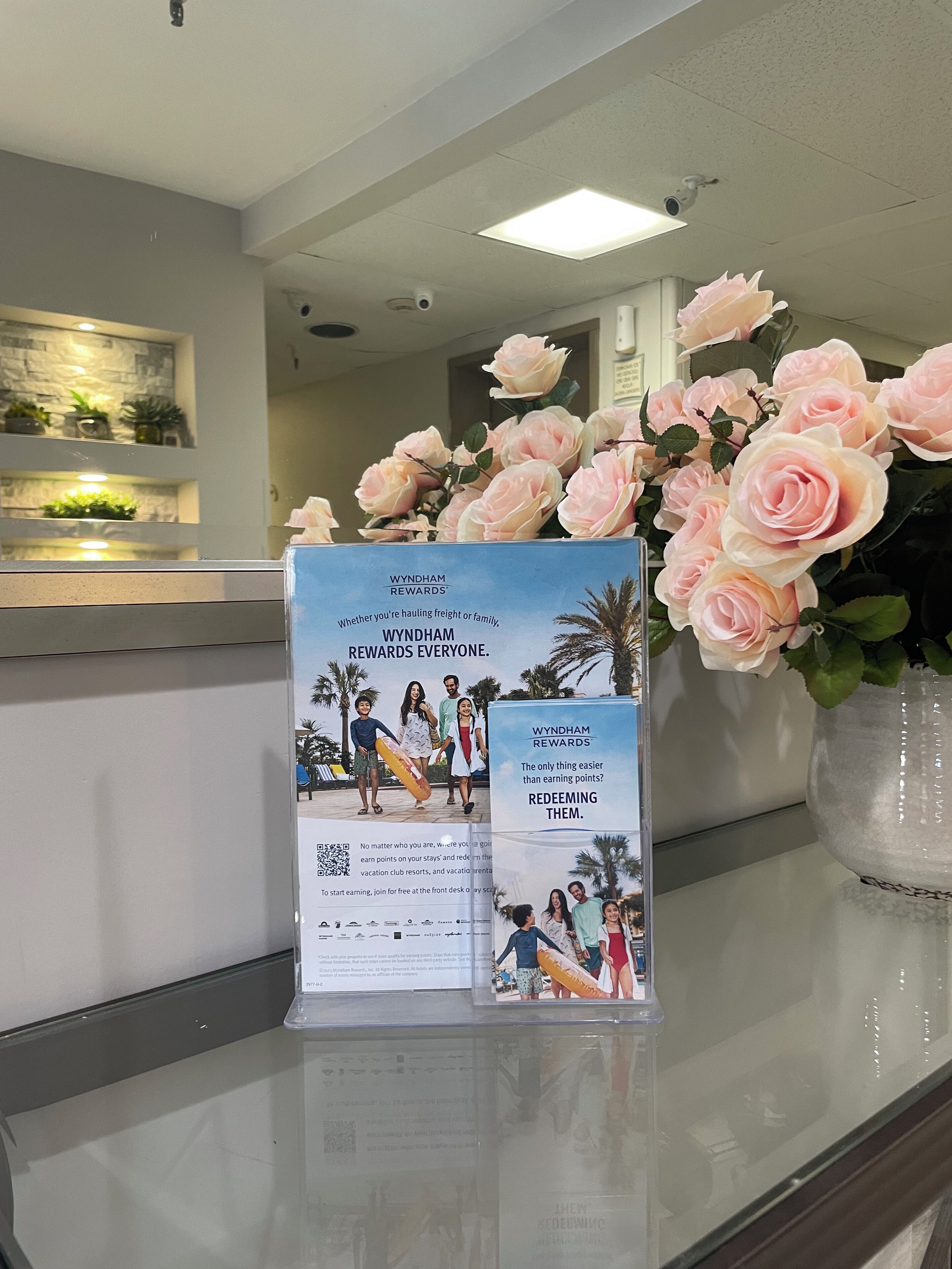 A reception desk with a Wyndham Rewards brochure, a vase of pink roses, and a modern interior with a stone wall and plants.