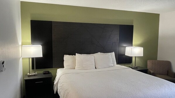 A hotel room with a large bed, two bedside tables with lamps, a chair, and a dark headboard.