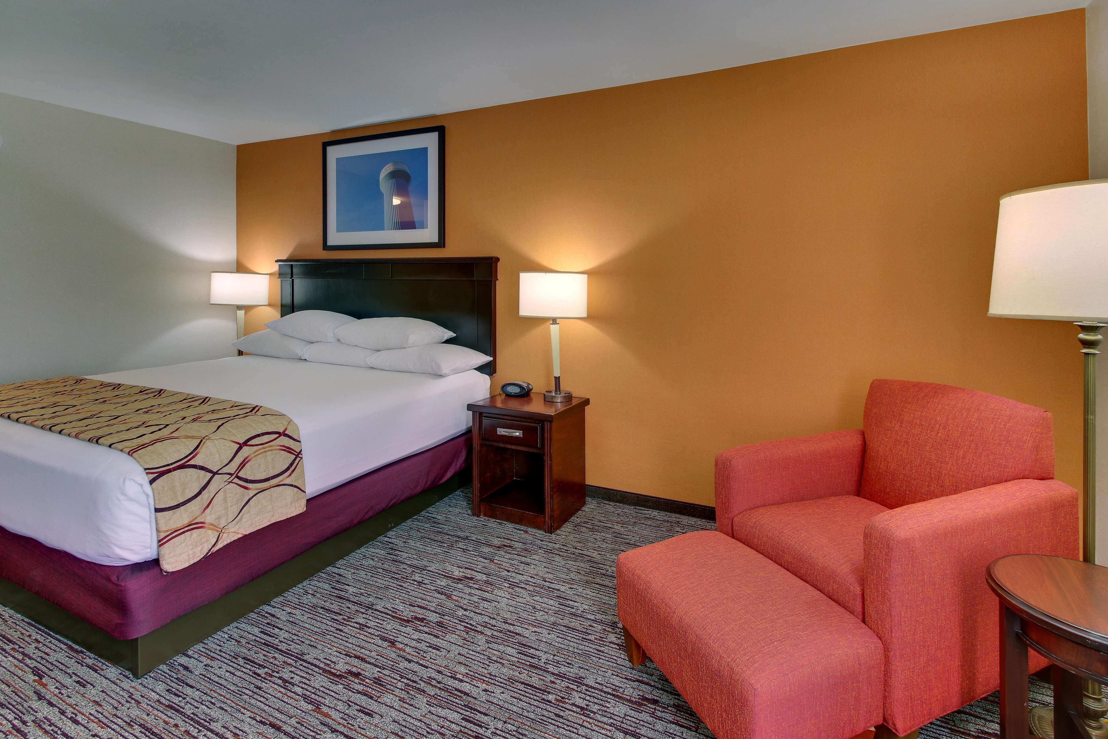 A hotel room with a bed, two bedside lamps, a red armchair, a small table, and a framed picture on the wall.