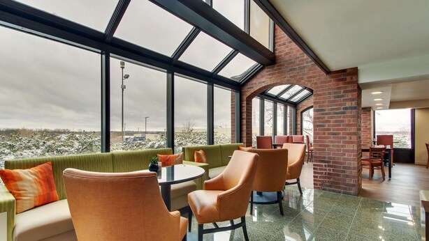 A modern interior with a high ceiling, large windows, and a brick wall. There are orange chairs and a green sofa with orange cushions. - Pear Tree Inn St. Louis Arnold (Arnold)