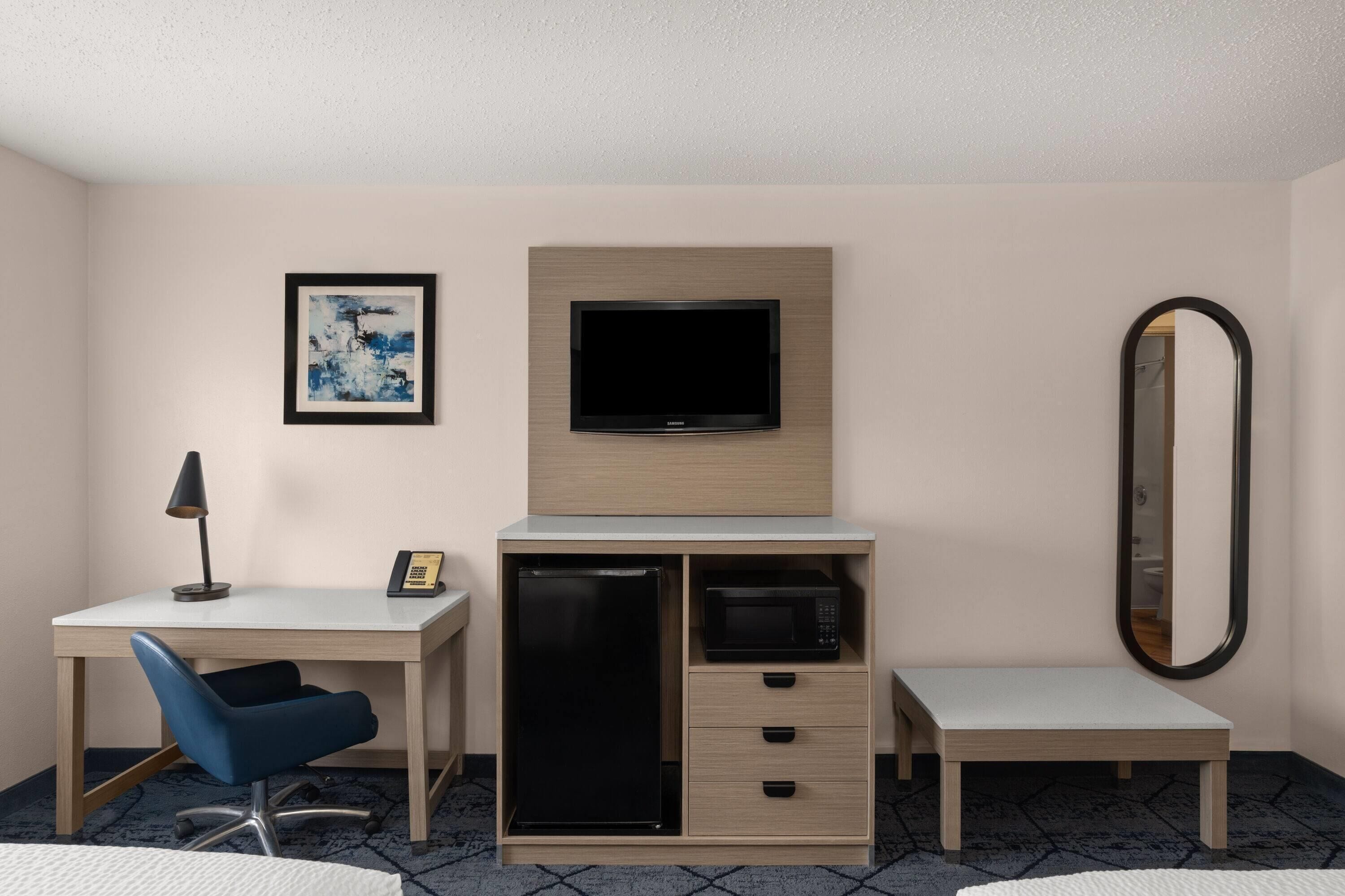 Habitación de hotel con escritorio, silla, televisor, microondas y un espejo grande.