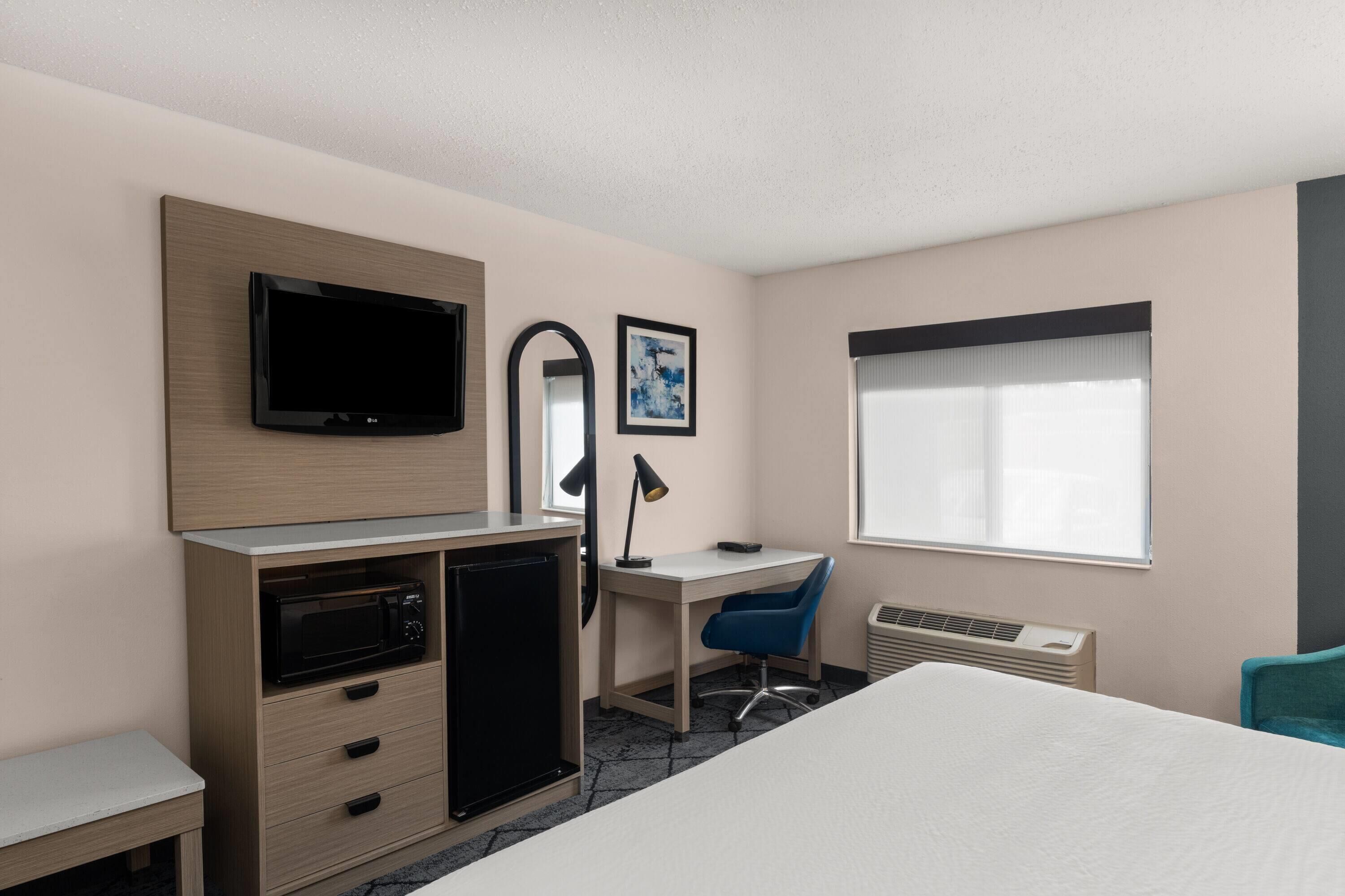 A hotel room with a bed, a desk, a television, a microwave, a heater, and a window with curtains.