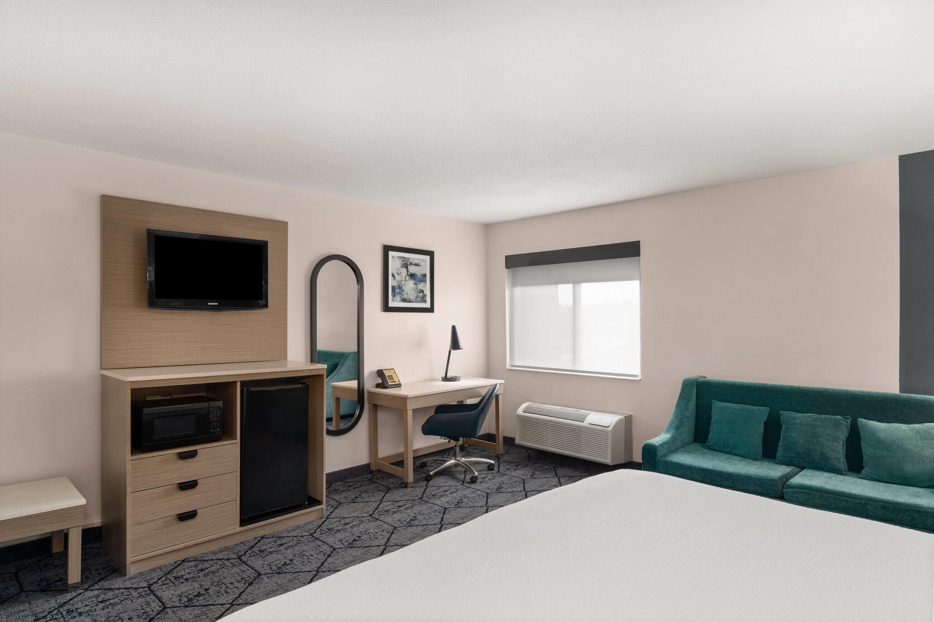 A hotel room with a bed, a desk, a chair, a television, a mirror, a radiator, and a window.