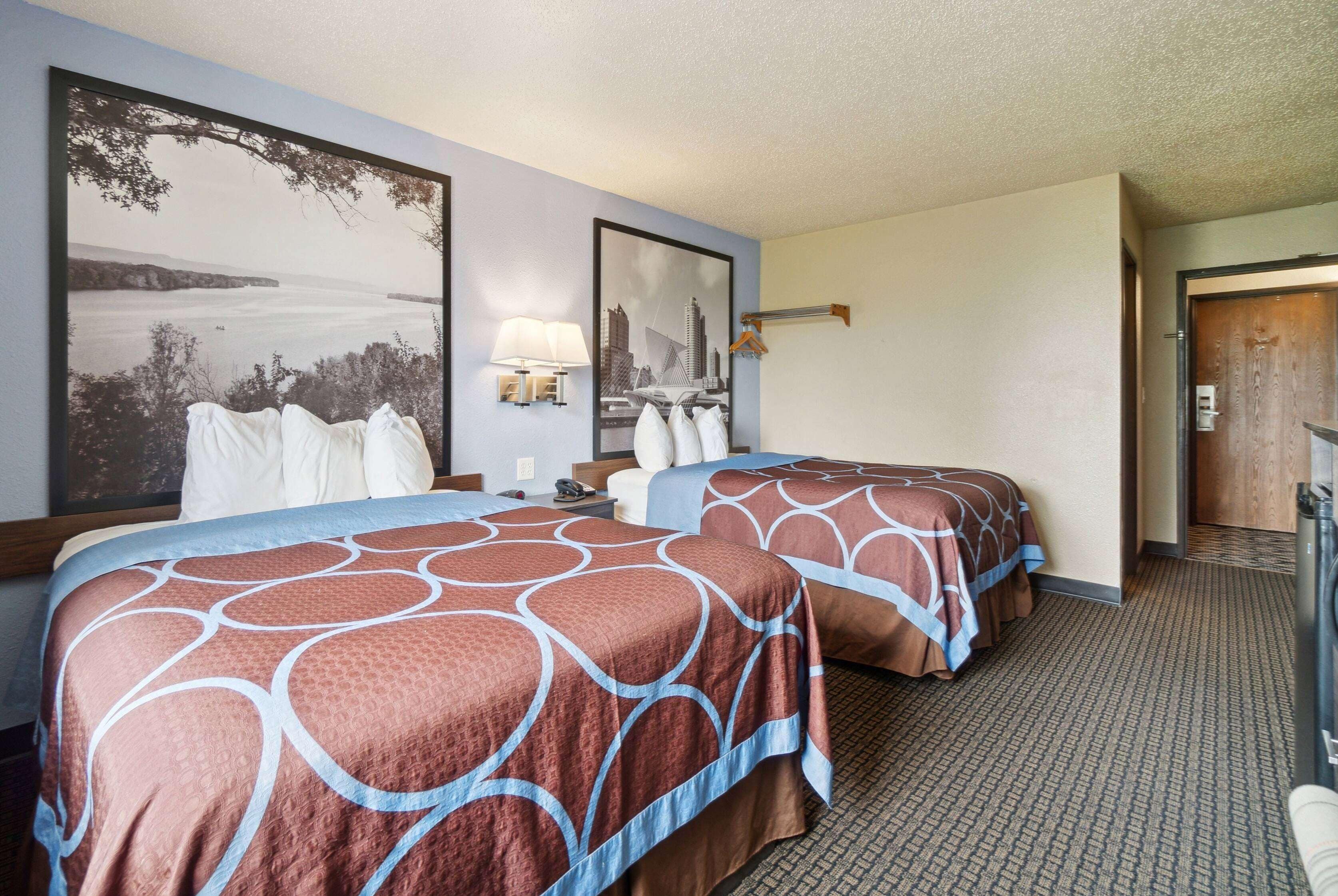 A hotel room with two beds, a cityscape painting, a lamp, and a door.
