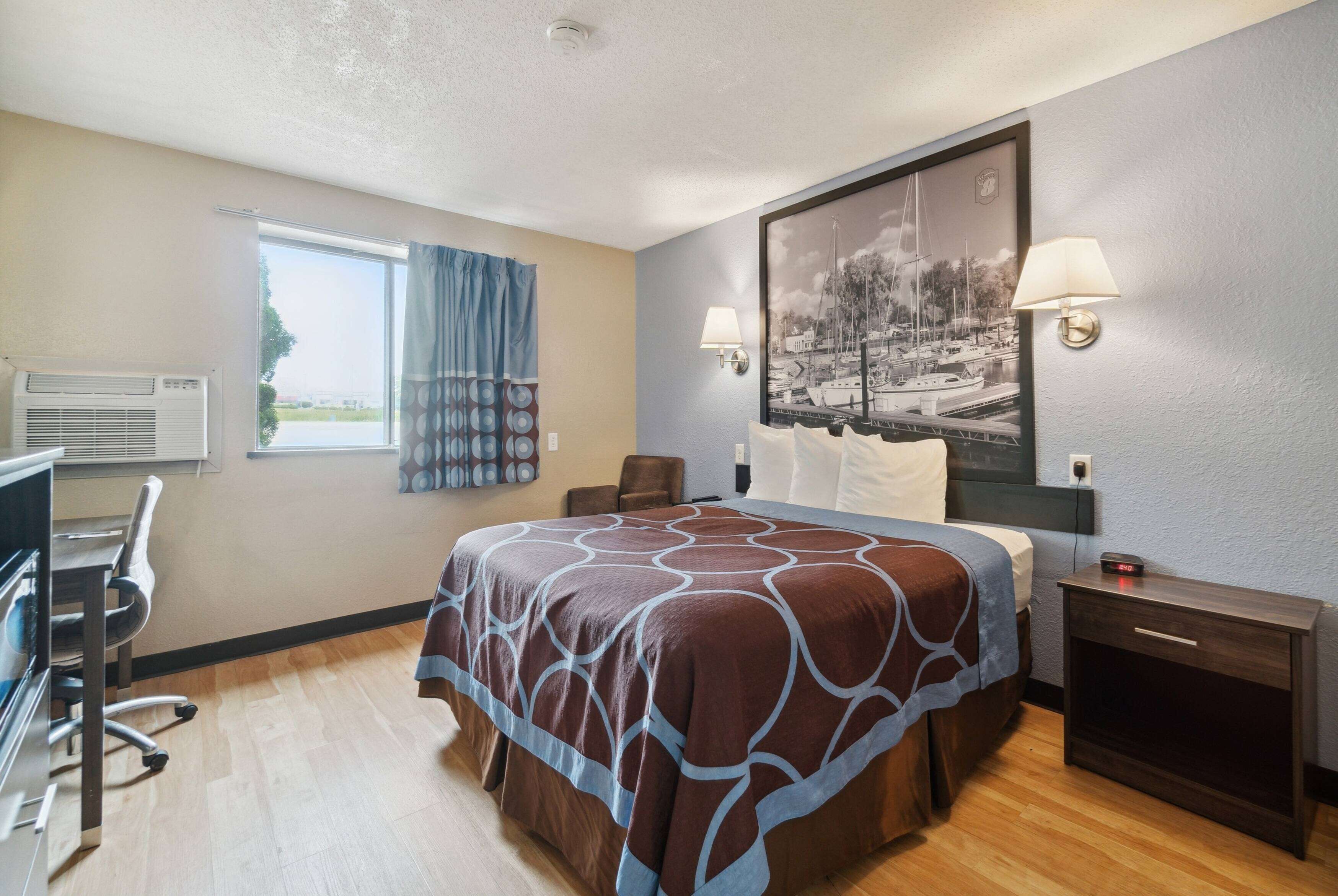 A hotel room with a large bed, a desk, an air conditioning unit, a nightstand, and a wall-mounted lamp.
