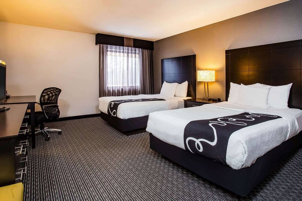 A hotel room with two beds, a desk, a chair, a lamp, and a large window with curtains.