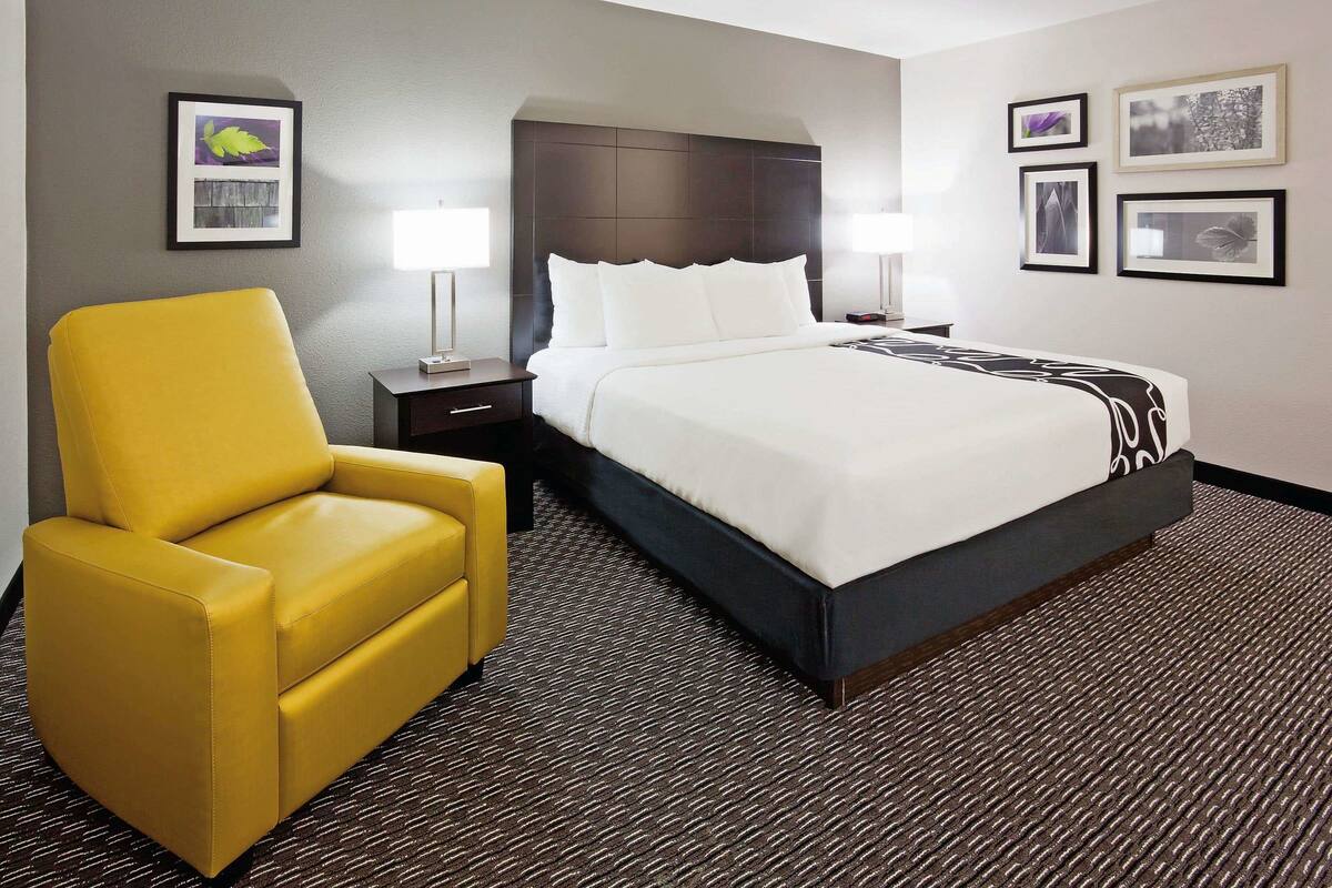 A hotel room with a large bed, a yellow armchair, two bedside tables with lamps, and framed pictures on the walls.