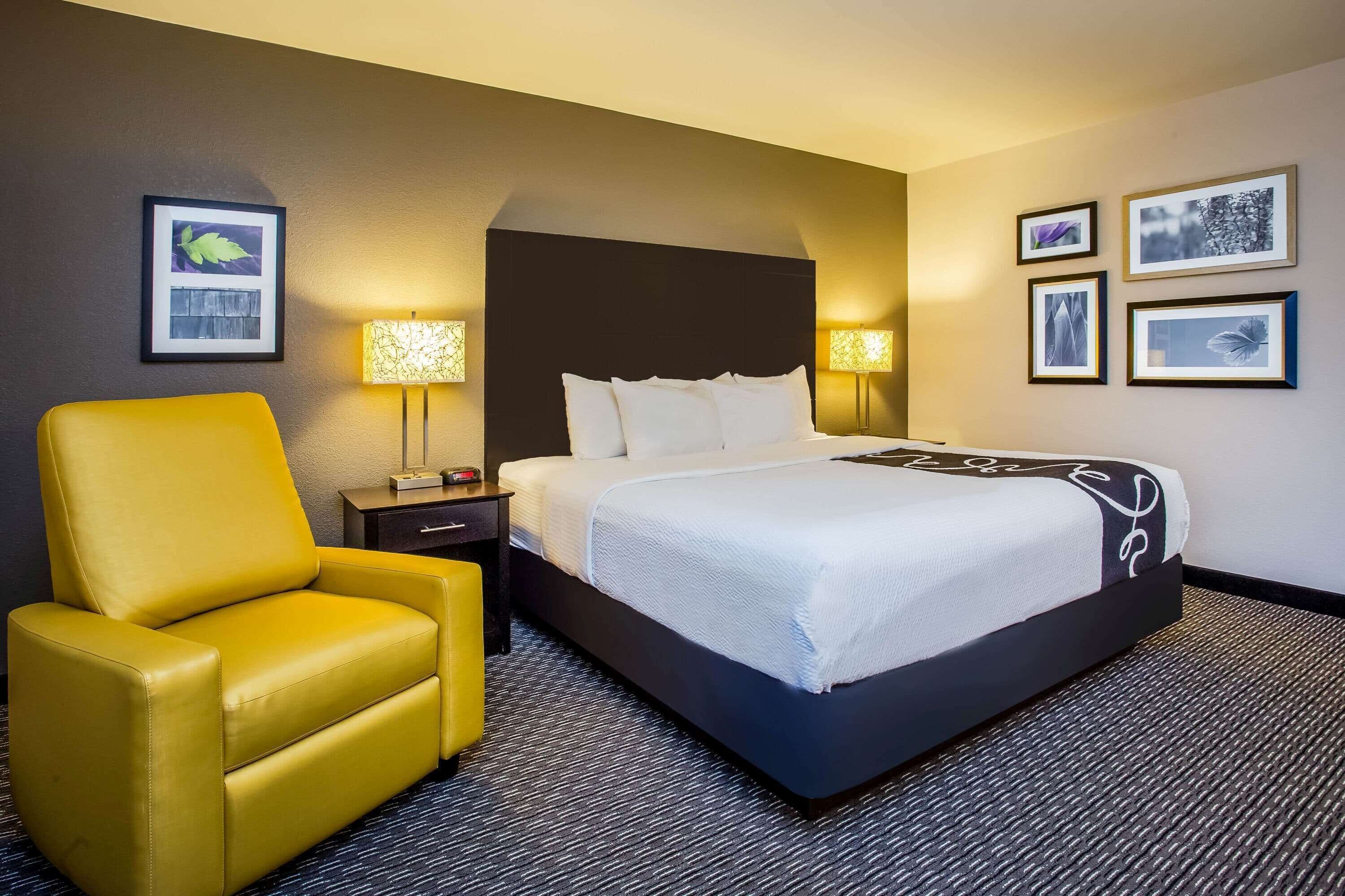 A hotel room with a large bed, a yellow armchair, a nightstand, and wall-mounted lamps.