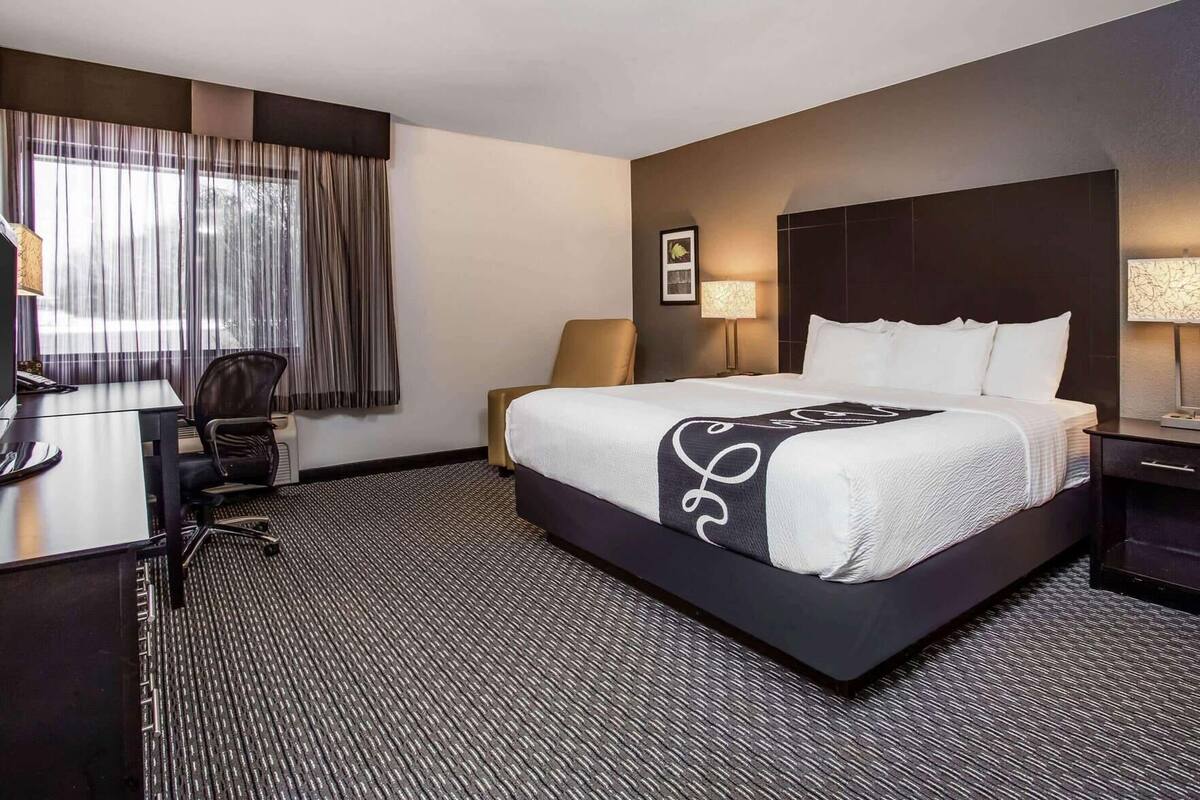 A hotel room with a large bed, a desk, a chair, a lamp, and a window with curtains.