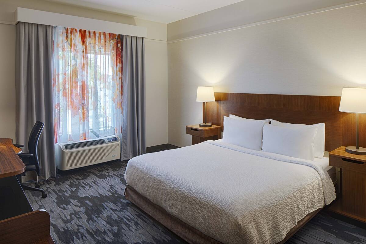 A hotel room with a large bed, two bedside tables with lamps, a desk, a chair, and a window with curtains.