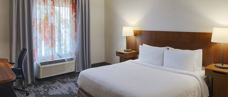 A hotel room with a large bed, two bedside tables with lamps, a desk, a chair, and a window with curtains.