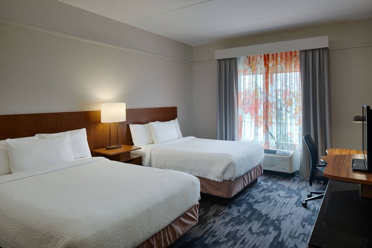A hotel room with two beds, a desk, a chair, a lamp, and a window with curtains.