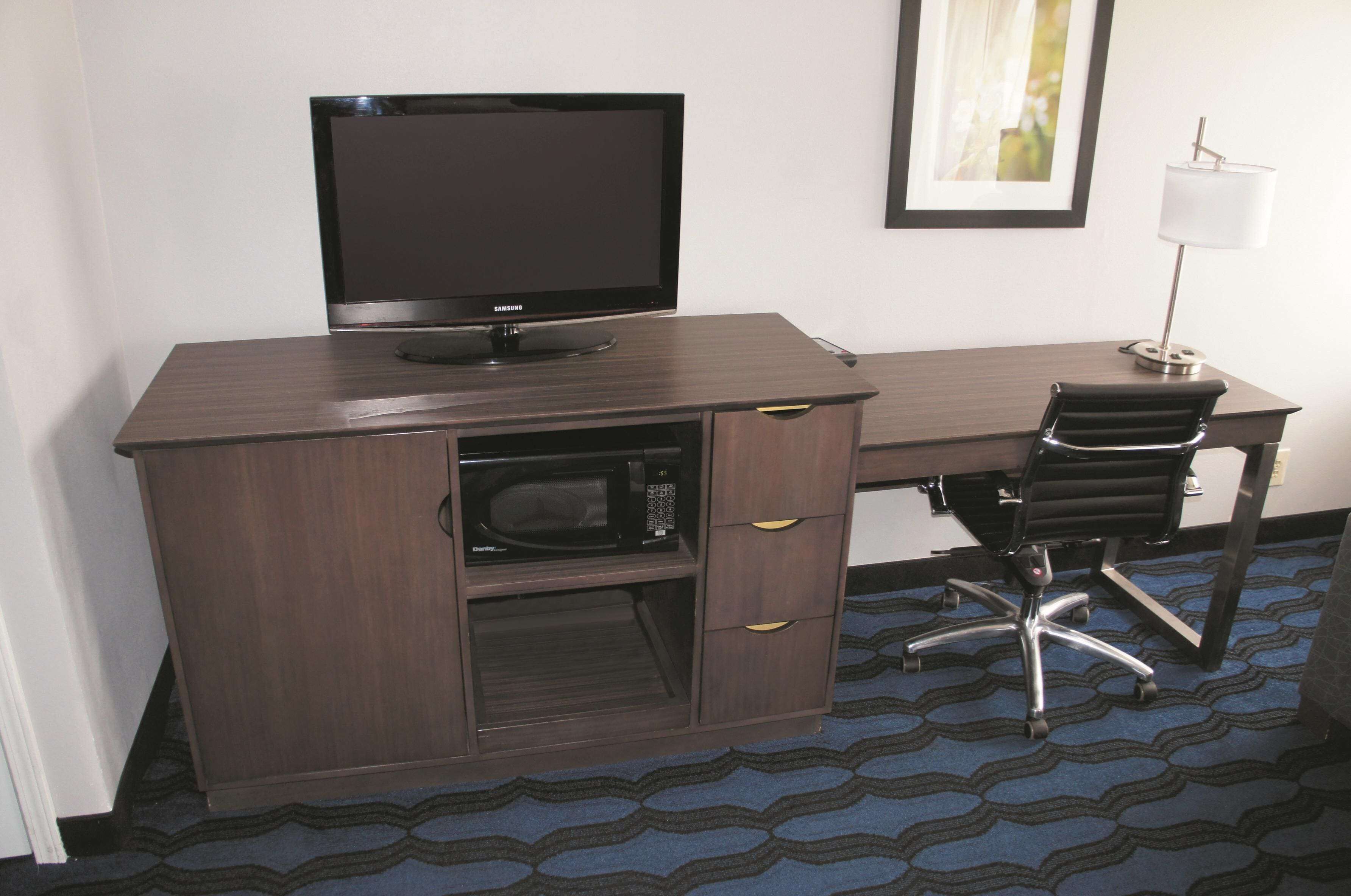 A hotel room with a dark wood desk, a flat-screen TV, a microwave, a black office chair, and a lamp.