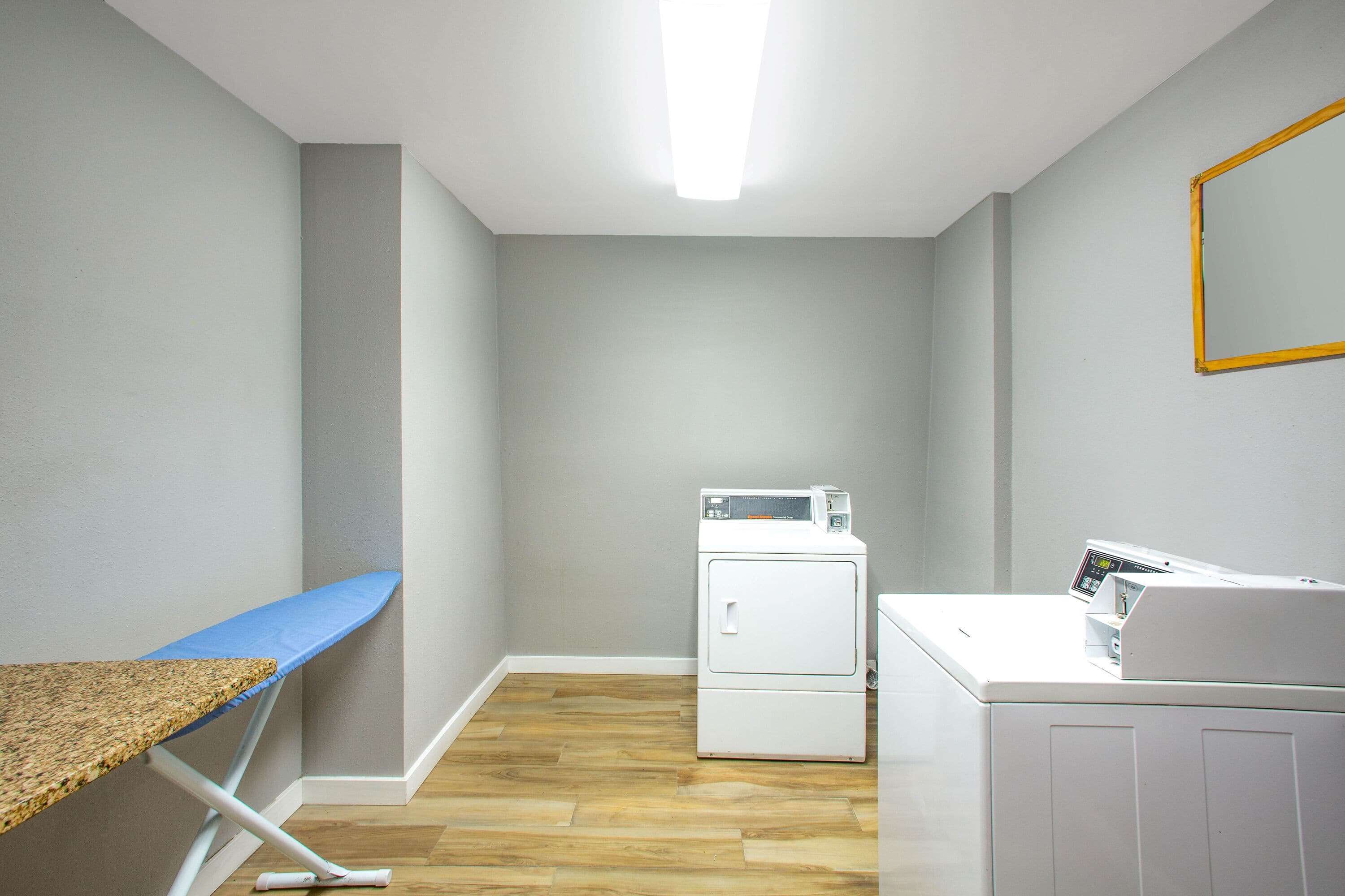 A laundry room with a washing machine, dryer, ironing board, and a mirror on the wall.