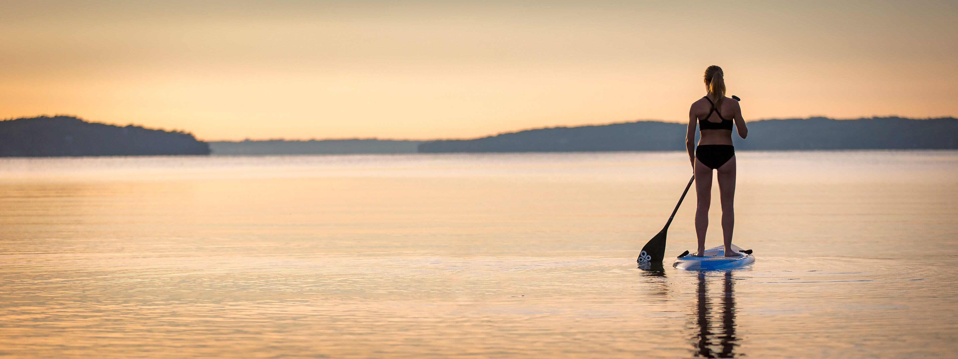 Une personne pratiquant le stand-up paddle sur une étendue d'eau calme au coucher du soleil.