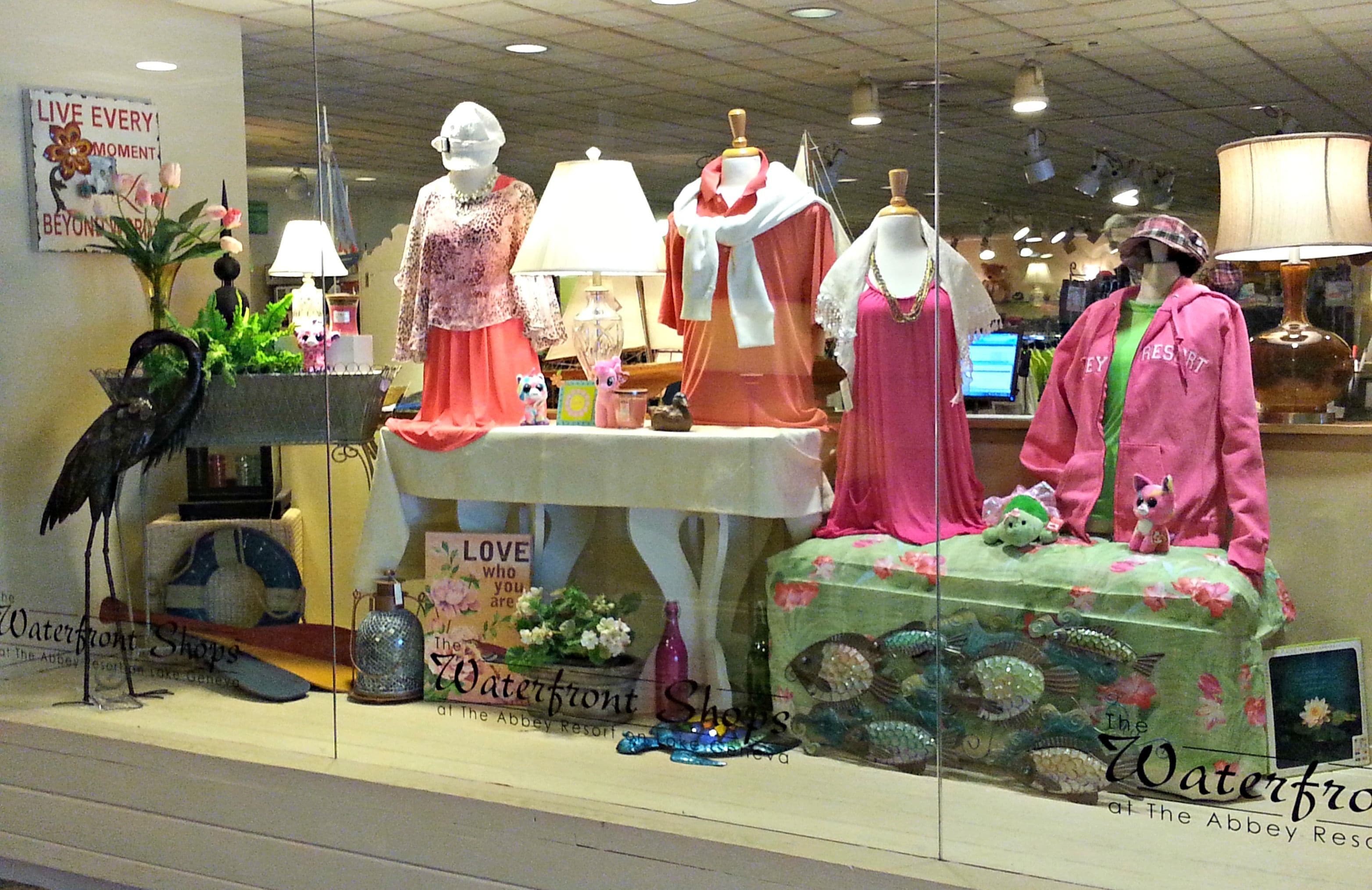 A store window display with mannequins dressed in various outfits, a sign that reads "Waterfront Shops at The Abbey Resort", and decorative items like a bird, flowers, and a fish tank.