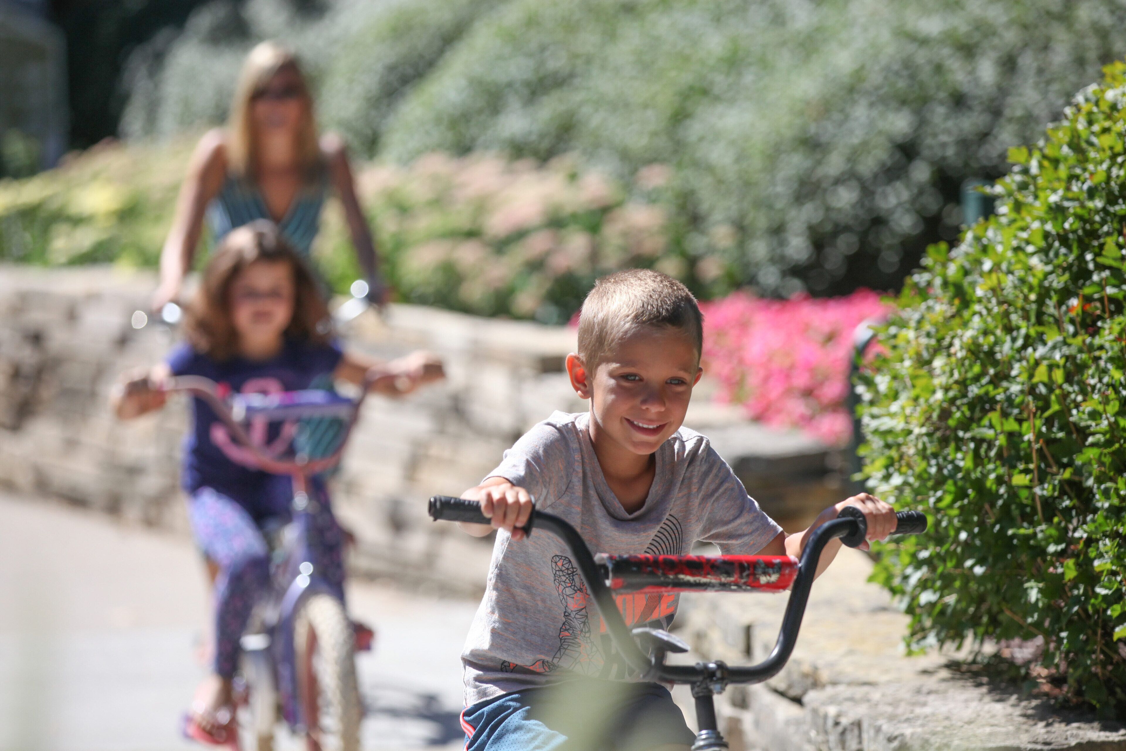 Niño montando en bicicleta por un camino pavimentado, con follaje y flores al fondo.