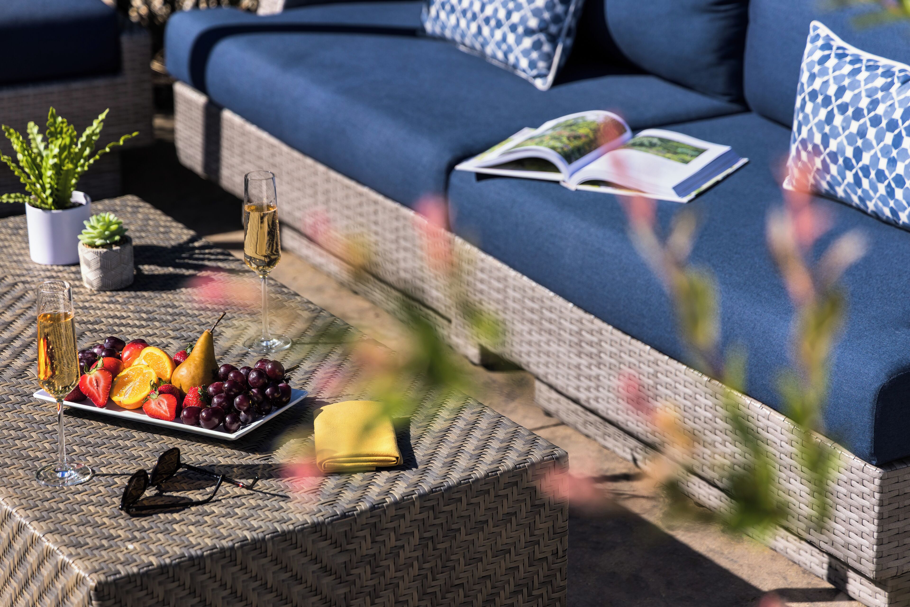 A wicker sofa with blue cushions, a glass of wine, and a fruit platter on a woven table.