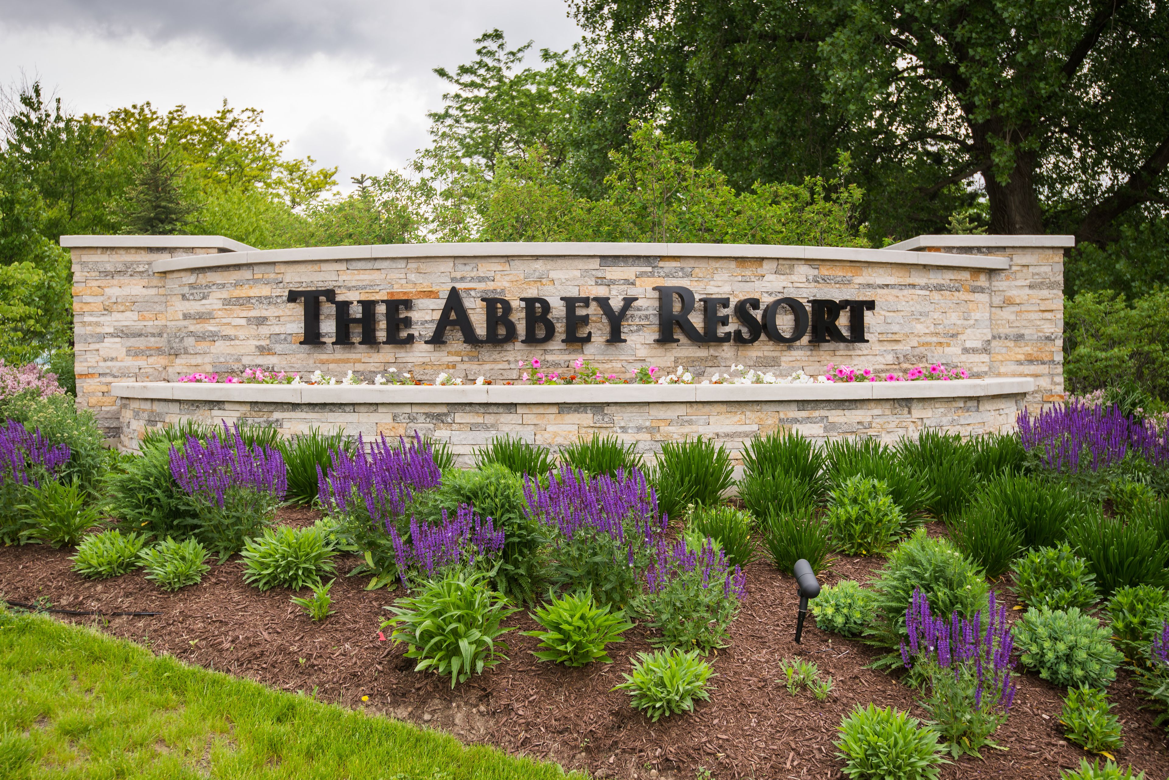 En stenskilte med teksten "The Abbey Resort", omgivet af en anlagt have med forskellige planter og blomster.