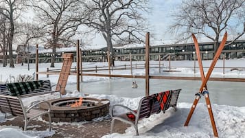 Un espacio exterior cubierto de nieve con un brasero, bancos y esquíes apoyados en un poste de madera.