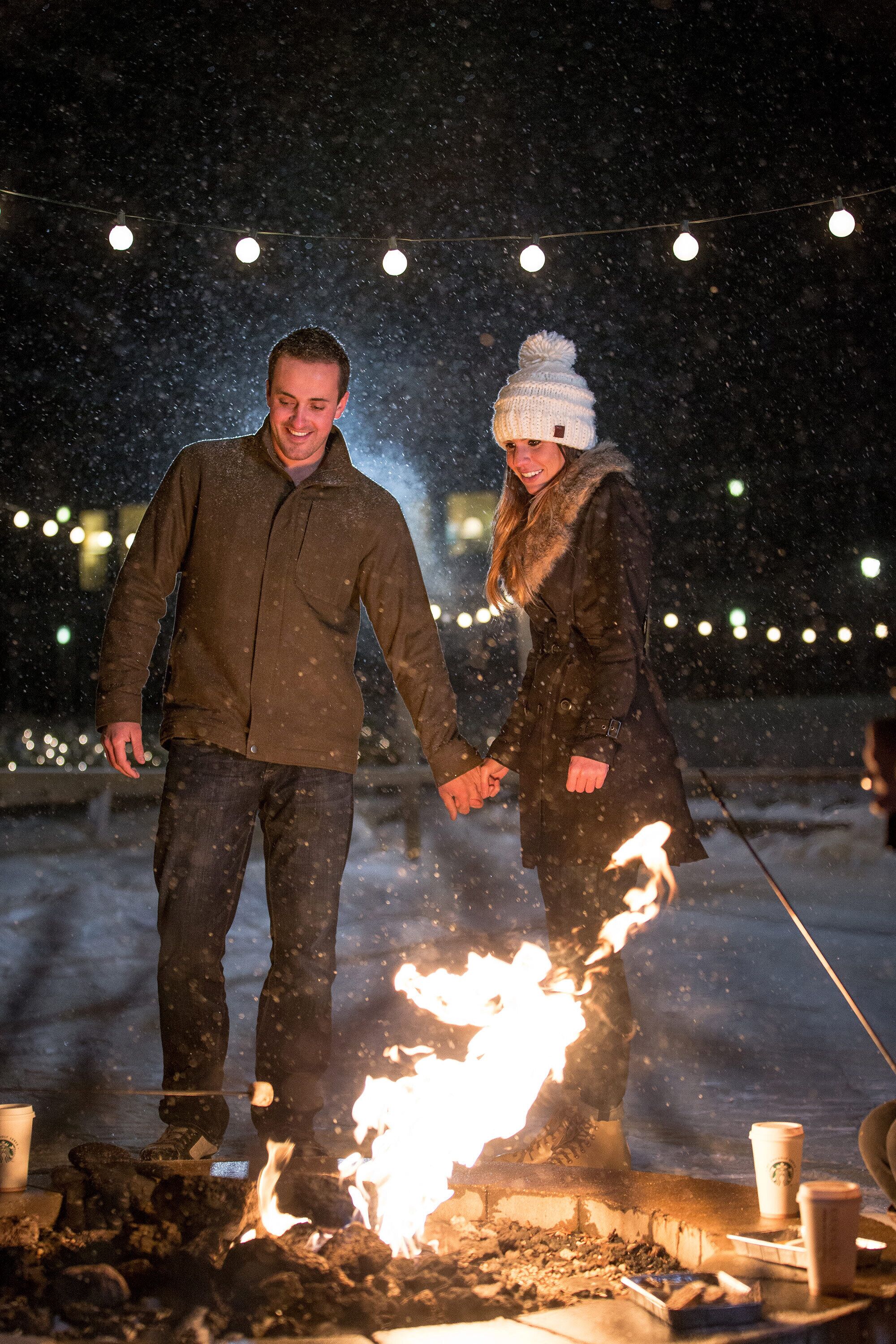 Twee personen die hand in hand staan bij een kampvuur, met lichtjes op de achtergrond.