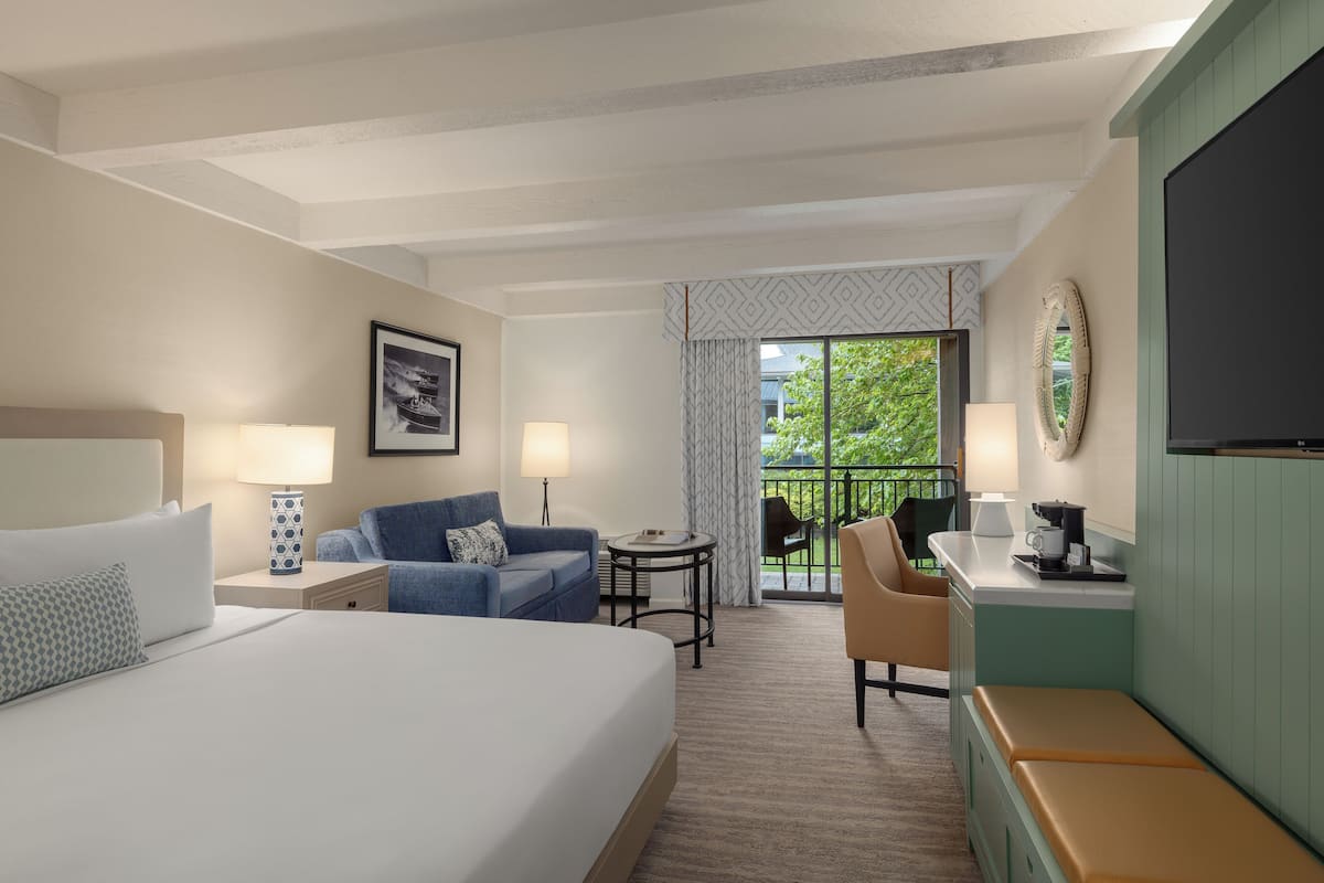 A hotel room with a large bed, a sofa, a small table, a chair, a TV, and a balcony with a view of greenery.