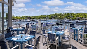Un espace extérieur aménagé pour les repas, avec des tables et des chaises, donnant sur une marina où sont amarrés des bateaux.