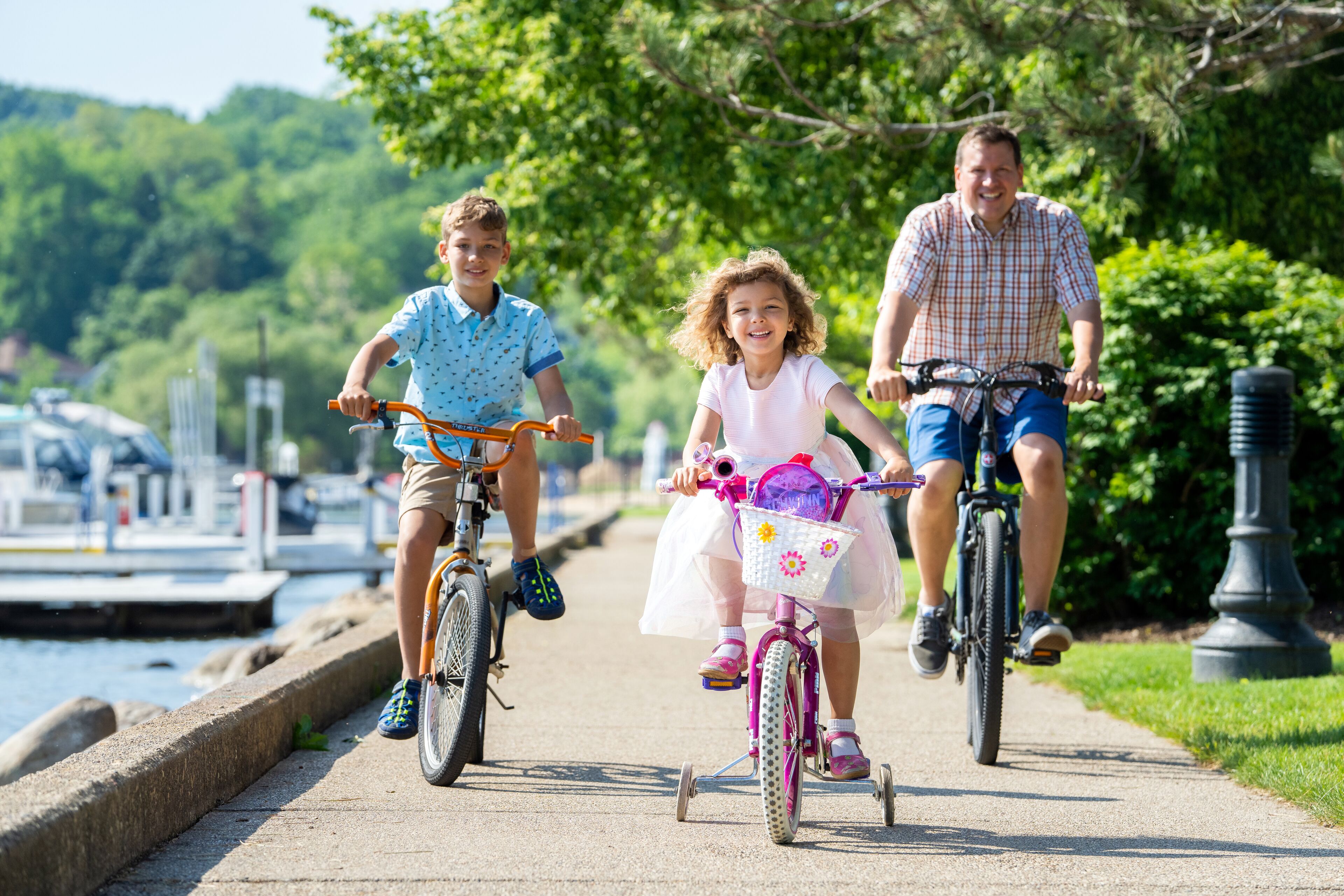 Three people riding bicycles on a paved path alongside a body of water, with greenery and a lamp post in the background.